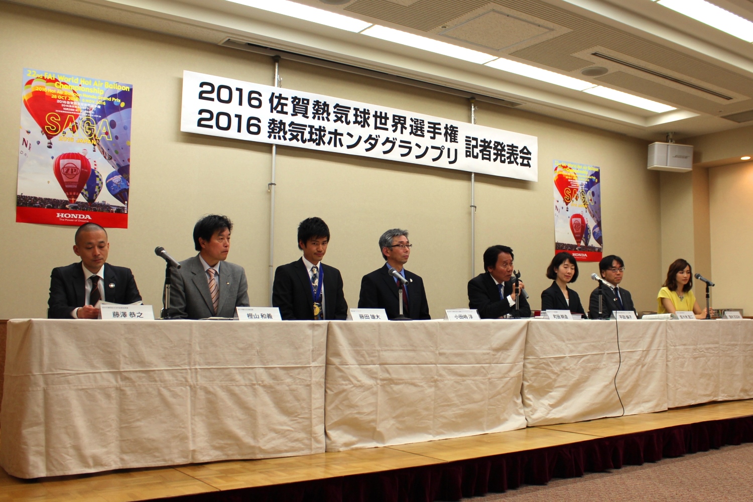 「2016佐賀熱気球世界選手権 2016熱気球ホンダグランプリ記者発表会」と書かれた横断幕が掲げられた記者発表会で、壇上の長いテーブルに並んだ優木まおみさんと7名の出席者がマイクを前に座り、背景にはバルーンが描かれたポスターが掲示されている、会見中の様子を捉えた写真