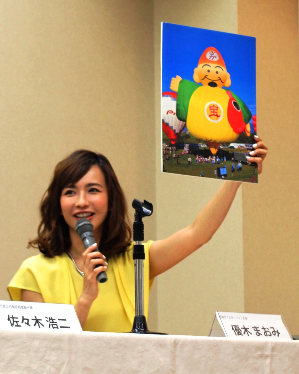 「2016佐賀熱気球世界選手権」の記者発表会にて、優木まおみさんがマイクを手に笑顔で話し、左手で恵比寿様の姿をした色鮮やかな恵比須バルーンが青空に浮かぶ写真を掲げながら、大会の魅力を紹介している様子を捉えた写真