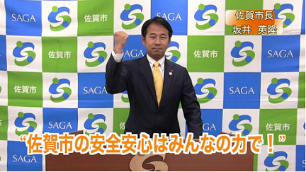佐賀市のロゴが並ぶ背景前でスーツ姿の市長が右手を握り拳にし、前方の台には「佐賀市の安全安心はみんなの力で!」という大きなオレンジ文字が表示された写真