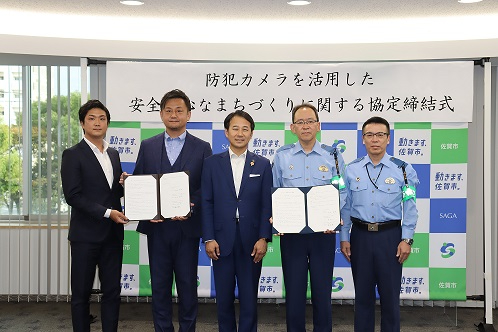 防犯カメラ活用の協定締結式で市長を中心にスーツ姿と制服姿の関係者が文書を掲げ並んで撮影された場面の写真
