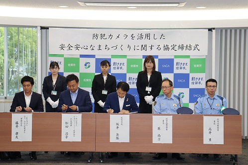 「防犯カメラを活用した安全安心なまちづくりに関する協定締結式」と書かれた横断幕の前で、スーツ姿の男性3名が書類に署名し、その傍らに警察官2名と案内役の女性3名が並んで立っている、締結式の様子を写した写真