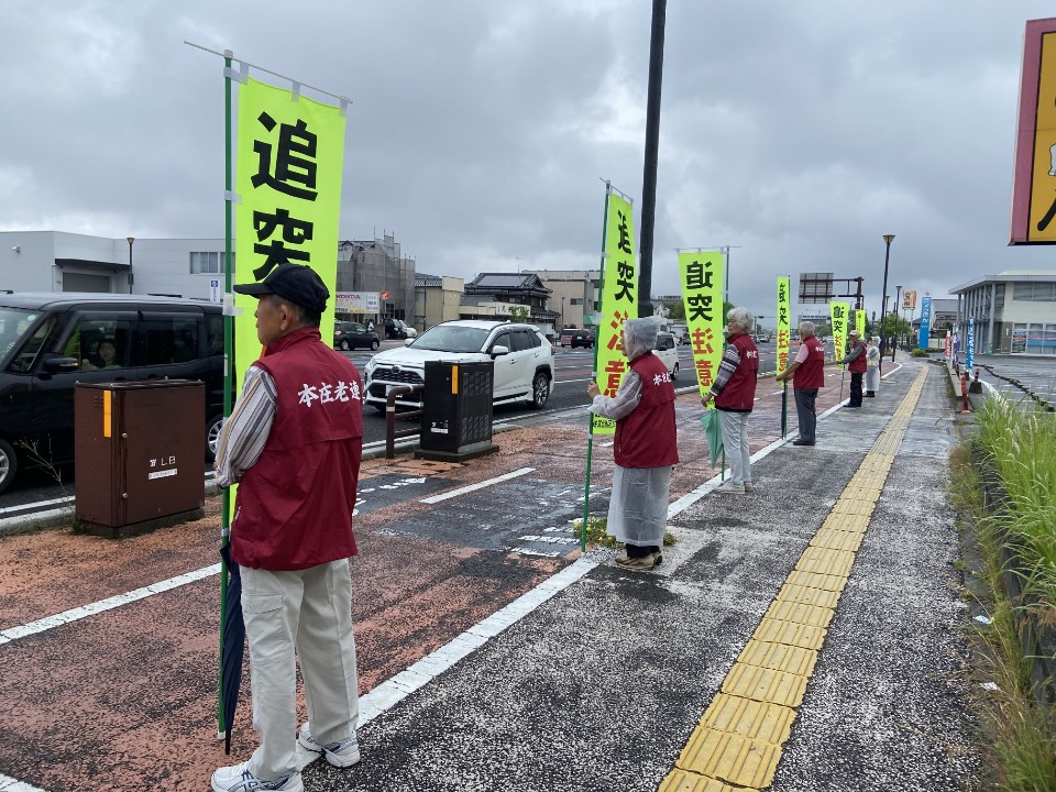 雨天の歩道に整然と並んだ赤いベストを着た参加者たちが、道路を走行する自動車を運転する人達に向けて黄色い旗を掲げ交通安全を呼びかけている様子の写真