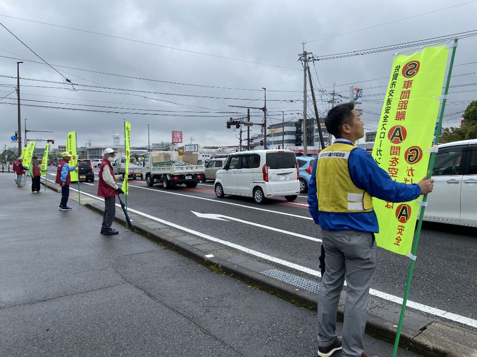 雨の道路沿いで参加者たちが車道脇に間隔を保って整列し、鮮やかな黄色い旗を掲げ交通安全を呼びかけている様子の写真
