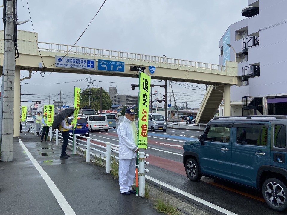 雨の道路沿いで参加者が黄色い交通安全旗を掲げ歩道に整然と並び、歩道橋前で通行車両に注意喚起を行っている写真