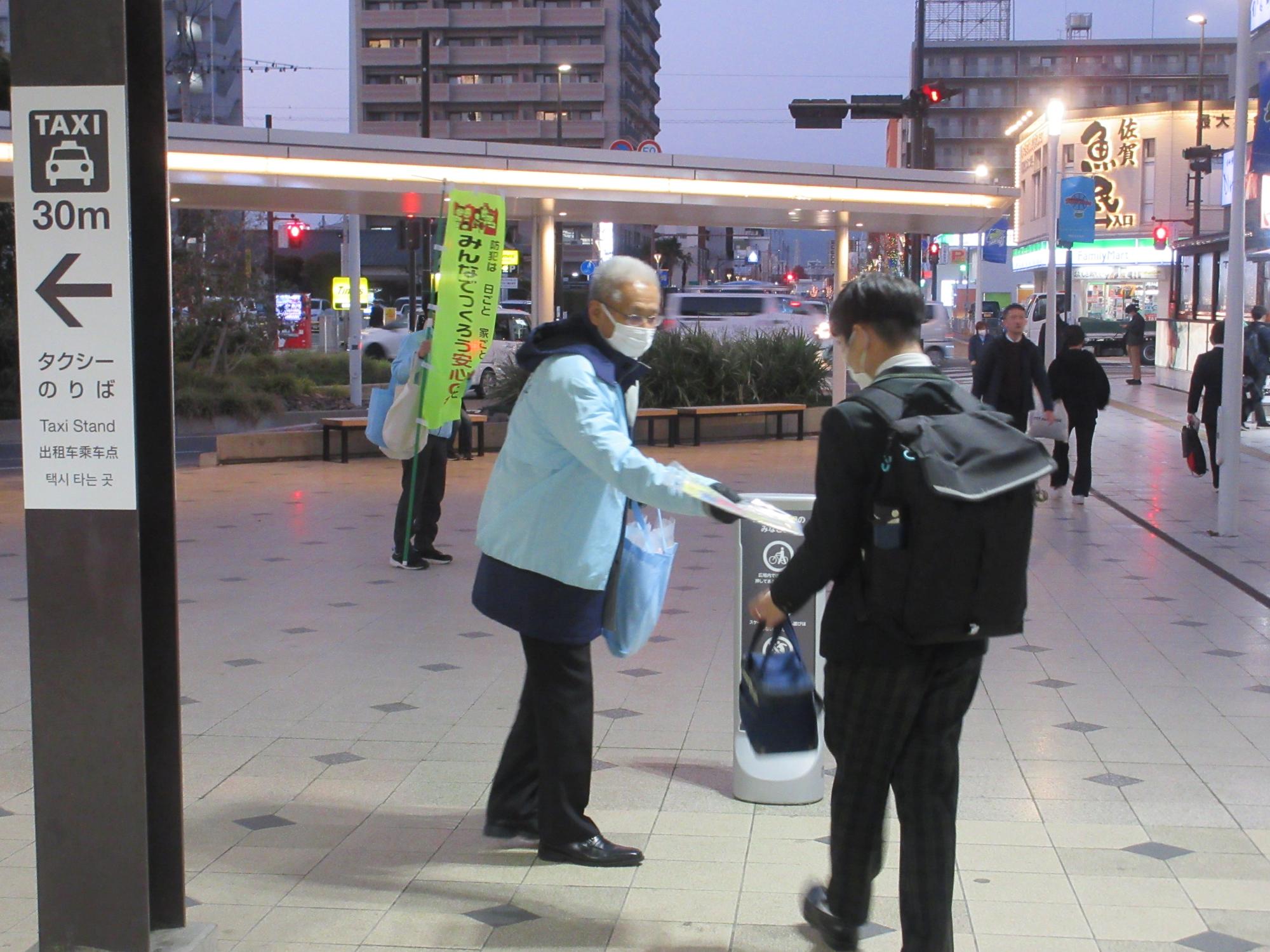 夕暮れ時の駅前広場にて、水色のジャンパーを着用した参加者の男性が、通りがかったリュックサック姿の学生に啓発チラシを手渡している、街頭キャンペーンの活動の様子を捉えた写真