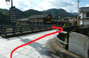 橋のたもとに石碑と欄干が並び、山間の住宅街へ向かう赤い矢印が道順を示している写真