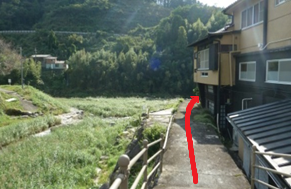 川沿いの細い舗道が民家の脇を緩やかに曲がり、進行方向を赤い矢印で明確に示して安全な通路を案内する様子の写真