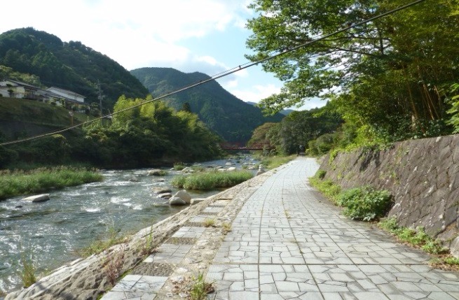 川沿いに石畳の遊歩道が緩やかに続き、山々と木々に囲まれた穏やかな風景が広がる写真
