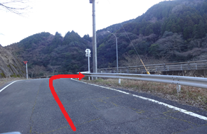 山間の舗装路がガードレール沿いに左へ曲がる進行方向を赤い矢印で示した写真