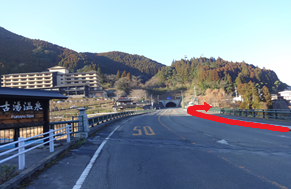 山間の温泉街へ続く広い道路が橋を渡った先で左へ曲がる方向を赤い矢印で示した写真
