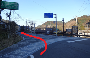 道路脇の案内板が立つ山間の交差点で左方向へ進む道筋を赤い矢印が明確に示している写真