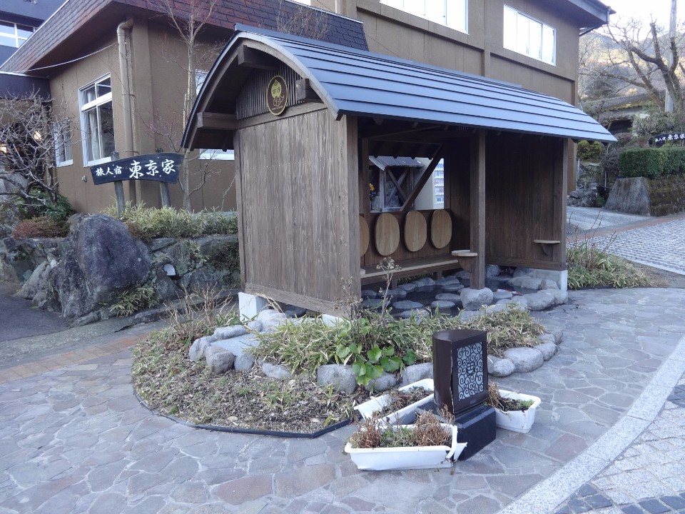 温泉街の建物前に木造の東屋が佇み、石畳の小道と植栽に囲まれた落ち着いた休憩空間を写した写真