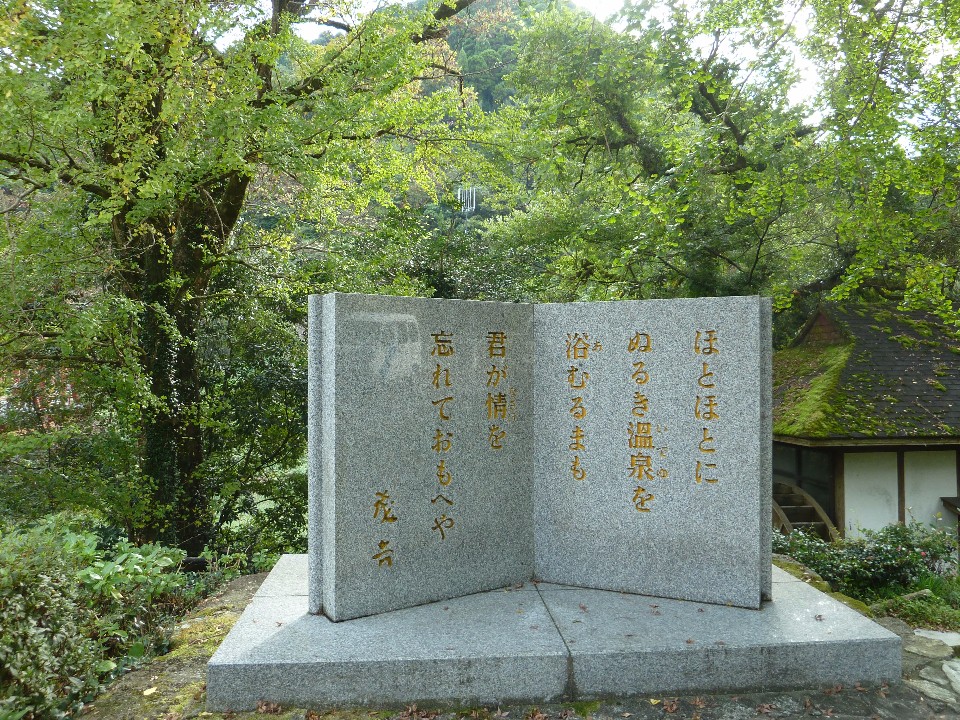 山間の緑豊かな木々に囲まれた場所に開いた本形の石碑が静かに佇み、表面に美しい金色の詩文が刻まれている写真
