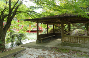 緑に包まれた東屋の奥に赤い橋がのぞき、静かな川辺の木陰に休憩できる空間が広がる写真