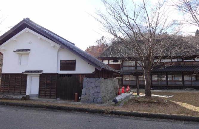 白壁の蔵風建物と木造の母屋が並び、冬木立の下に石垣や庭が広がる静かな和風景観の写真