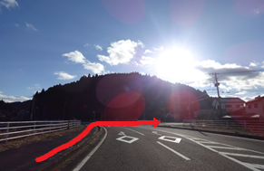 強い逆光に照らされた山沿いの道路が白いガードレールに沿って右へ進む方向を赤い矢印で示した写真