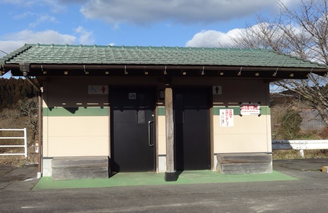 緑色の瓦屋根を載せた公衆トイレ棟が正面に並び、男女と多目的用の黒い扉が日差しを受けて明瞭に見える写真