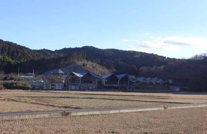 広い枯れ田の向こうに森と青空が続き、山裾にやまびこの湯の建物が見える写真