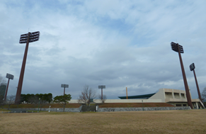 曇り空の下、広大な芝生広場の向こう側に野球場が位置し、空に向かって高くそびえ立つ複数の巨大な夜間照明塔が周囲を囲むように配置された、開放感と力強さが共存するみどりの森県営球場の写真