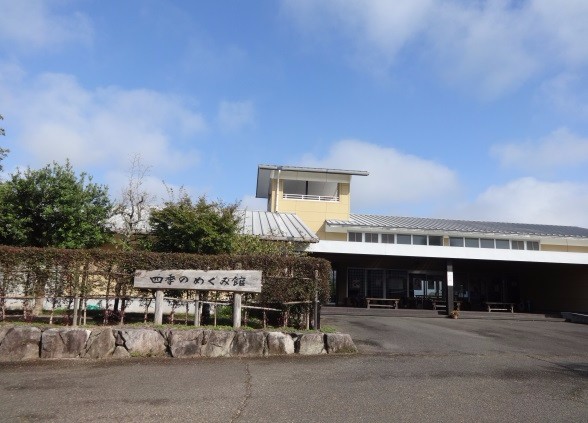 青い空と白い雲の下に建つ「四季のめぐみ館」という木製看板が掲げられた平屋建ての施設で、石垣と生け垣に囲まれたエントランス付近には舗装された道路が広がっている四季のめぐみ館の外観写真