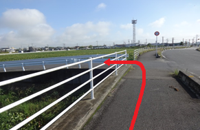左側に白い柵がある歩道を直進し、途中で左に曲がって橋のような場所を渡るルートが赤い矢印で示された、広々とした緑地や遠くの鉄塔を背景に、道順を視覚的にガイドする役割を持つ写真