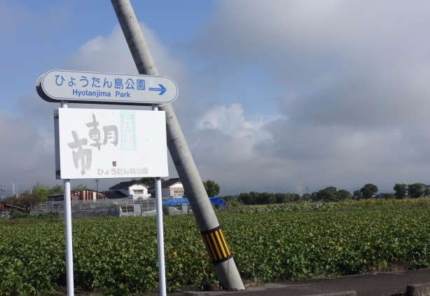 青空の下に広がる畑の脇に、「ひょうたん島公園」への方向を示す矢印付きの看板と「朝市」と書かれた白い看板が立っており、背景には遠くの山々や民家が薄い雲とともに写し出されている、のどかな屋外の風景を記録した写真