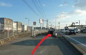 左側に大型トラックが並ぶフェンス沿いの広い歩道を直進し、前方の地下通路のようなスロープを下りるルートが赤い矢印で示された道順の写真