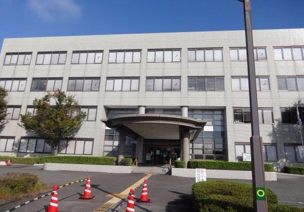 青空の下に建つグレーのタイル貼りの4階建てほほえみ館の正面の外観で、円形の庇がある玄関前には赤と白のカラーコーンが並び、建物の手前には緑の植栽と舗装された道路が広がっている、ほほえみ館入り口周辺の写真