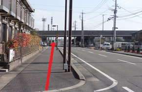 左側にアパートが並ぶ歩道を直進し、前方の高架下をくぐり抜ける進行方向が赤い矢印で示された、住宅街を通る平坦な車道と並行する歩道の風景から道順を説明する、視覚的な案内としての役割を持つ写真
