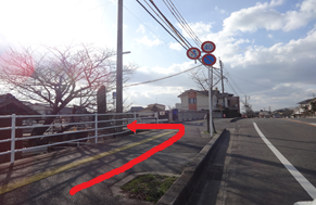 緩やかなカーブを描く道路から左側の歩道へ向かう赤い矢印が描かれ、白いガードレールの奥には冬の木々や石碑が立ち、右上には制限速度や駐車禁止の交通標識が並ぶ、住宅街の中の分岐点を案内する写真