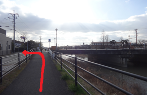 川沿いの遊歩道において、手前から進んできた経路から左側の建物が並ぶ道へと曲がる赤い矢印が描かれ、右側には川に架かる橋がある、サイクリングロードのルート分岐を案内する写真