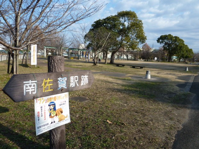 左側に「南佐賀駅跡」と書かれた木製の矢印型看板が立ち、その下にはマナー啓発のポスターが貼られ、奥にはベンチや木々が点在する広々とした南佐賀公園の写真