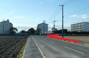 澄み渡る青空の下、田畑に囲まれた二車線の道路において、前方の交差点を右へと曲がり、その先に建つ数棟の集合住宅方面へと進むルートを、路面に合成された大きな赤い矢印で分かりやすく示した風景の写真