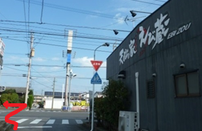 澄み渡る青空の下、黒い外壁に大きく「えび蔵」と店名が書かれた建物の角から、前方の横断歩道がある交差点を左へと曲がる進路を、赤い矢印で分かりやすく示したルート案内の写真
