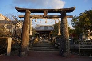 夕暮れ時の柔らかな光に照らされた神社において、注連縄が飾られた重厚な石造りの鳥居が立ち、その奥へと続く参道の先に瓦屋根の門や境内の木々が佇む、静かで厳かな本庄神社の写真