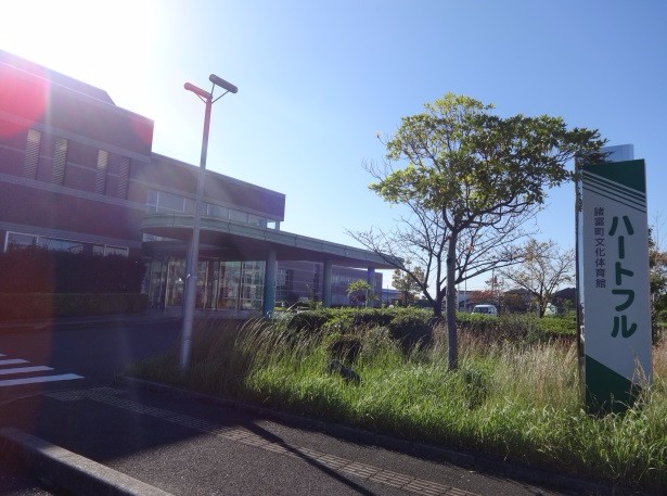 青空の下、近代的な赤レンガ調の建物の前に「ハートフル 諸富町文化体育館」と書かれた大きな緑色の看板が立ち、手前には秋の気配を感じさせるススキが風に揺れる、施設の入り口付近の風景を捉えた写真