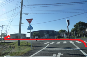 青空が広がる快晴の下、横断歩道のある交差点を捉え、電柱や交通標識が立ち並ぶ道路の先に低層の建物が見え、進路を示すような赤い矢印が左方向へと描かれた、日常的な街角の風景を捉えた写真