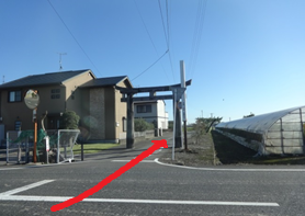 青空が広がる快晴の下、住宅地を通る道路の先に建つコンクリート造りの鳥居を捉え、その手前を右折して鳥居をくぐる進路を示す赤い矢印が描き込まれた、街角の風景を捉えた写真