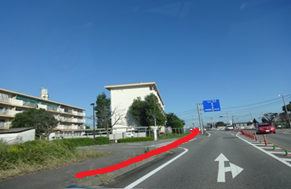 青空が広がる快晴の下、団地やアパートが立ち並ぶエリアの交差点を捉え、本道から左側の脇道へと進入する進路を示す赤い矢印が描き込まれた、見通しの良い郊外の風景を捉えた写真