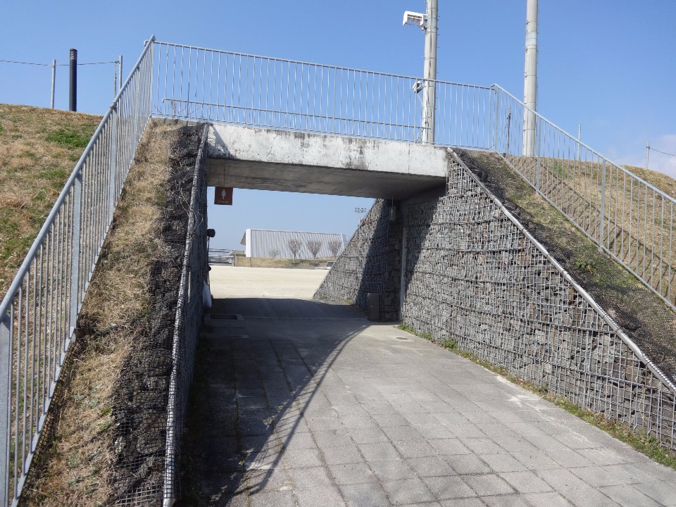 芝生が広がる堤防の下を通るコンクリート製のトンネルのような通路があり、側面は石を網で囲った蛇籠の壁面で構成され、通路の奥には広場が見え、上部には金属製のフェンスが設置された、晴天の日の公園内の写真