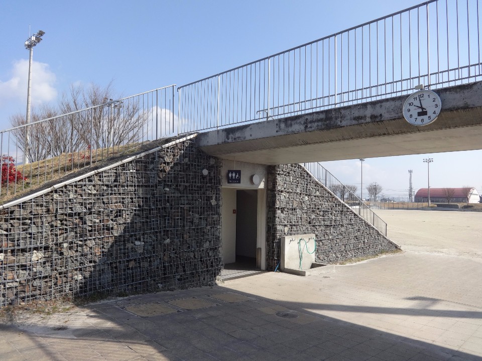 芝生の斜面の下にあるコンクリート通路の入り口付近を捉えており、通路の側面は網で石を固定した蛇籠で造られ、入り口の上にはトイレのマーク、通路の梁には丸時計が設置され、奥には広々とした運動場が見える写真