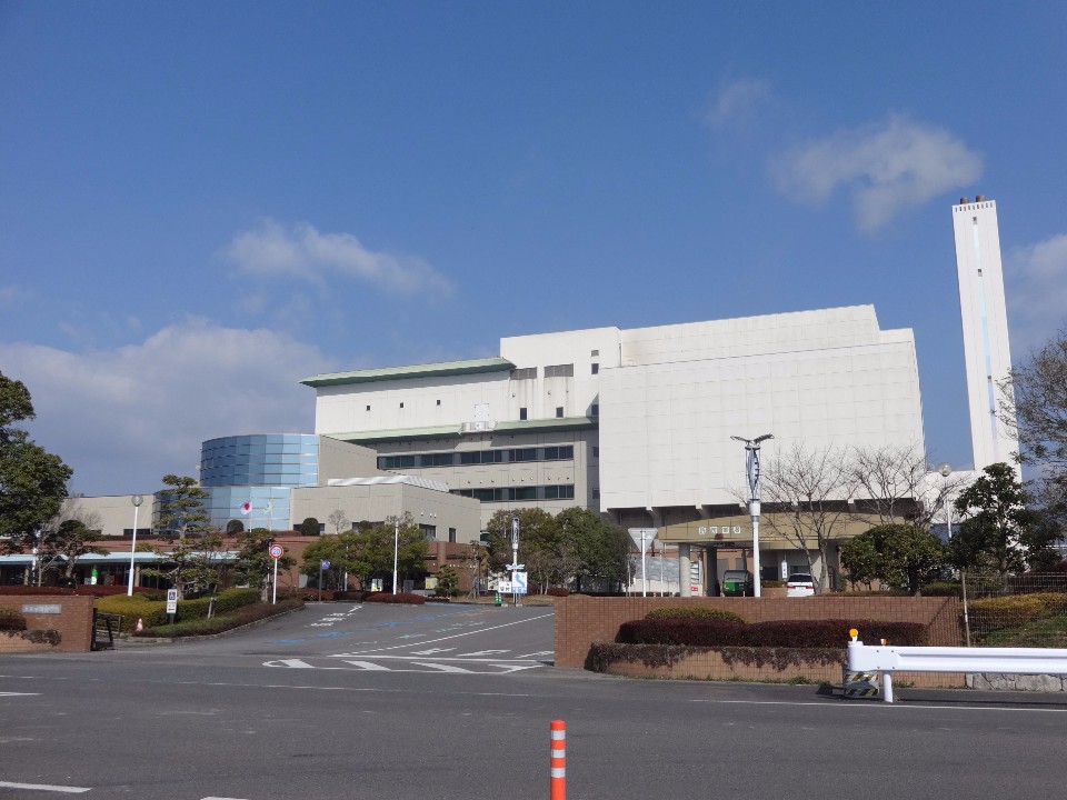 巨大な白い箱型の建物と細長い煙突が特徴的な施設の外観を捉えており、手前には植栽で整えられたアプローチ道路や駐車場、背後には青空が広がる、機能的で大規模な佐賀市エコプラザの景観を映し出した写真