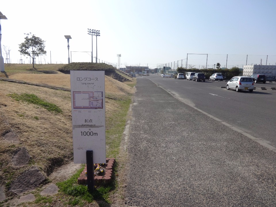 芝生の斜面と舗装された道路の境界に、「ロングコース 起点 1000メートル」と書かれたコース案内板が立っており、背景には駐車場に並ぶ車やスポーツ施設の照明塔、広々とした空が広がる、運動公園内の写真