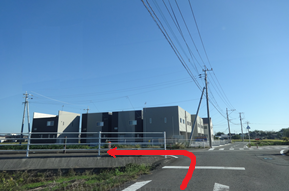 青空が広がる快晴の下、田園地帯の交差点を捉え、ガードレールの手前を左折して住宅が並ぶ方向へと進む赤い矢印が描き込まれた、見晴らしの良い郊外の風景を捉えた写真