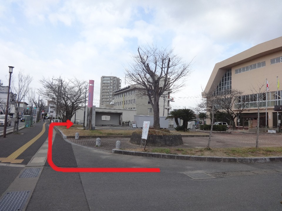 曇り空の下、舗装された道路を直進してから右折し、樹木や電柱が並ぶ歩道沿いを進むルートが赤い矢印で示され、右側に体育館のような大きなベージュ色の建物が建つ開けた広場付近の道順を視覚的にガイドする役割を持つ写真