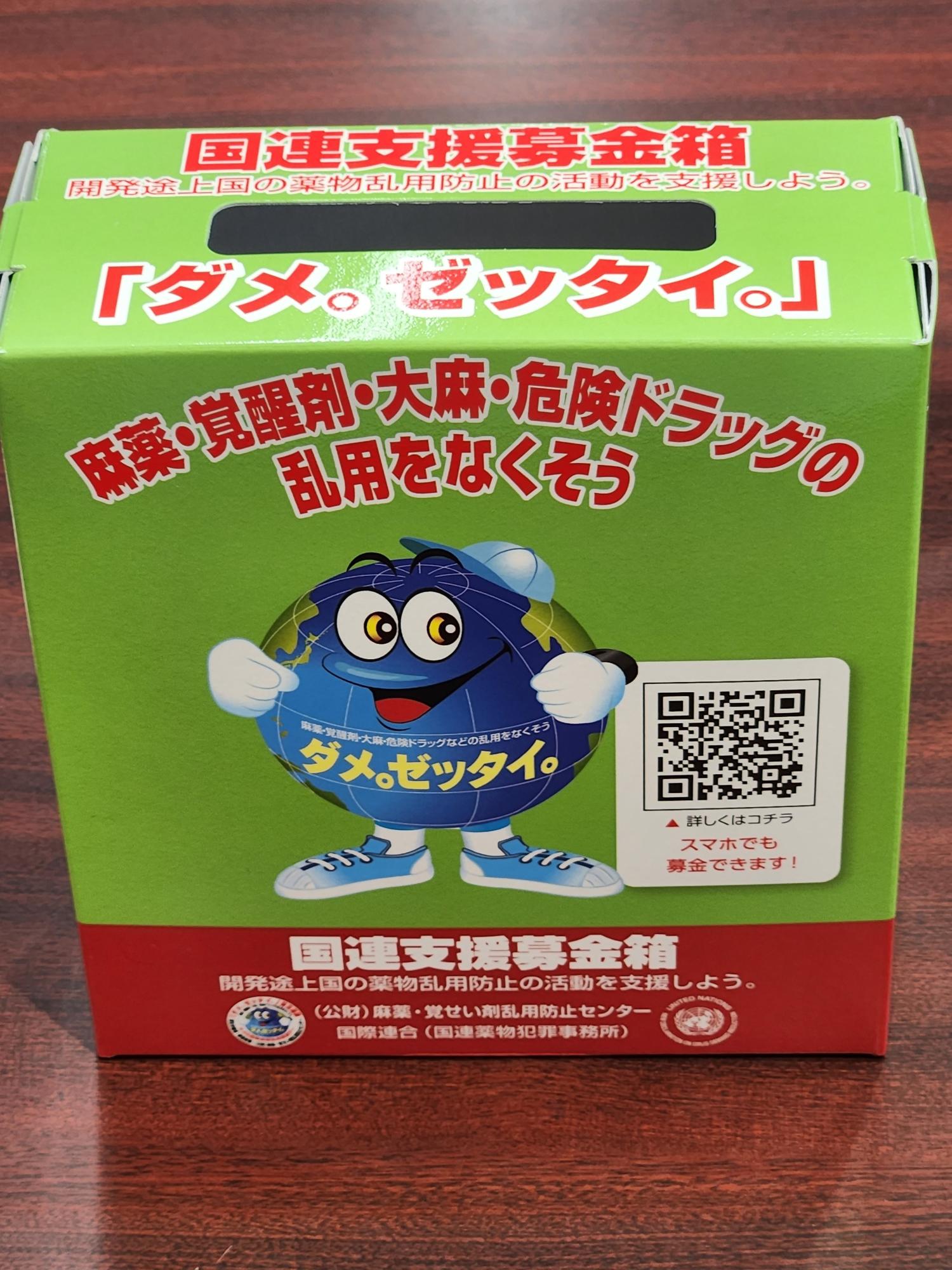 緑色の箱に「ダメ。ゼッタイ。」と赤文字で書かれ、麻薬・覚醒剤・大麻・危険ドラッグの乱用防止を訴えるメッセージと、青いキャラクターのイラスト、さらにQRコードと「国連支援募金箱」の文字が印刷された薬物乱用防止啓発用募金箱の写真