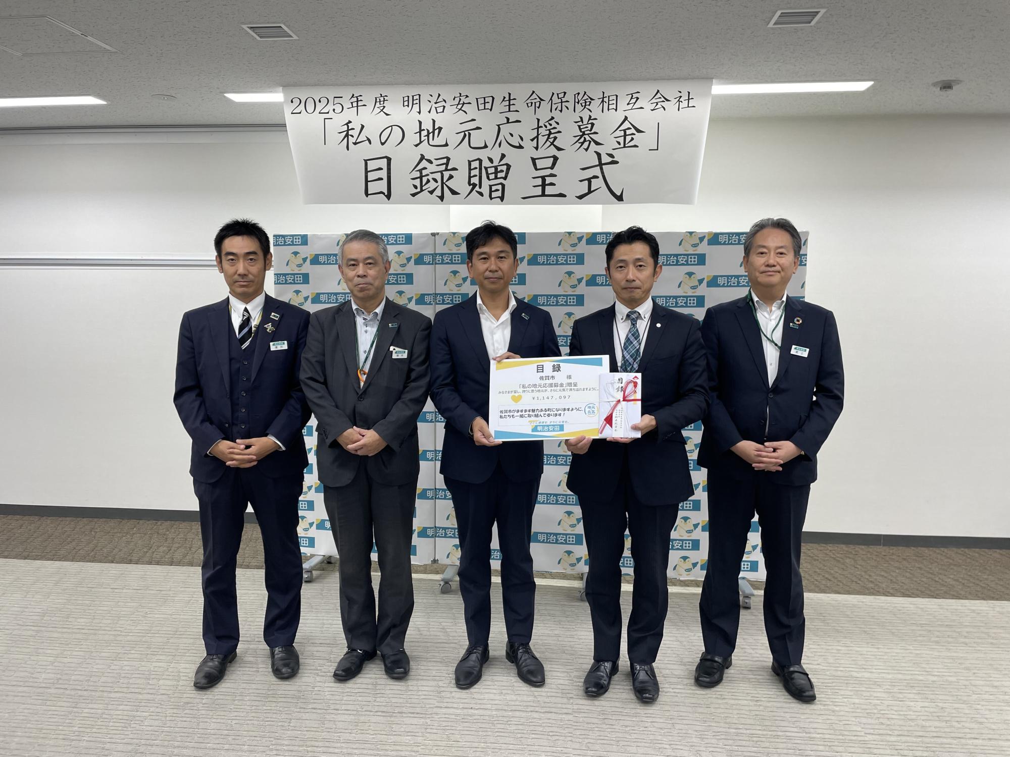 横断幕に「2025年度 明治安田生命保険相互会社 私の地元応援募金 目録贈呈式」と書かれた背景の前で、スーツ姿の5人が並び、中央の2人が大きな目録と熨斗付きの封筒を持っている贈呈式の様子を示す写真