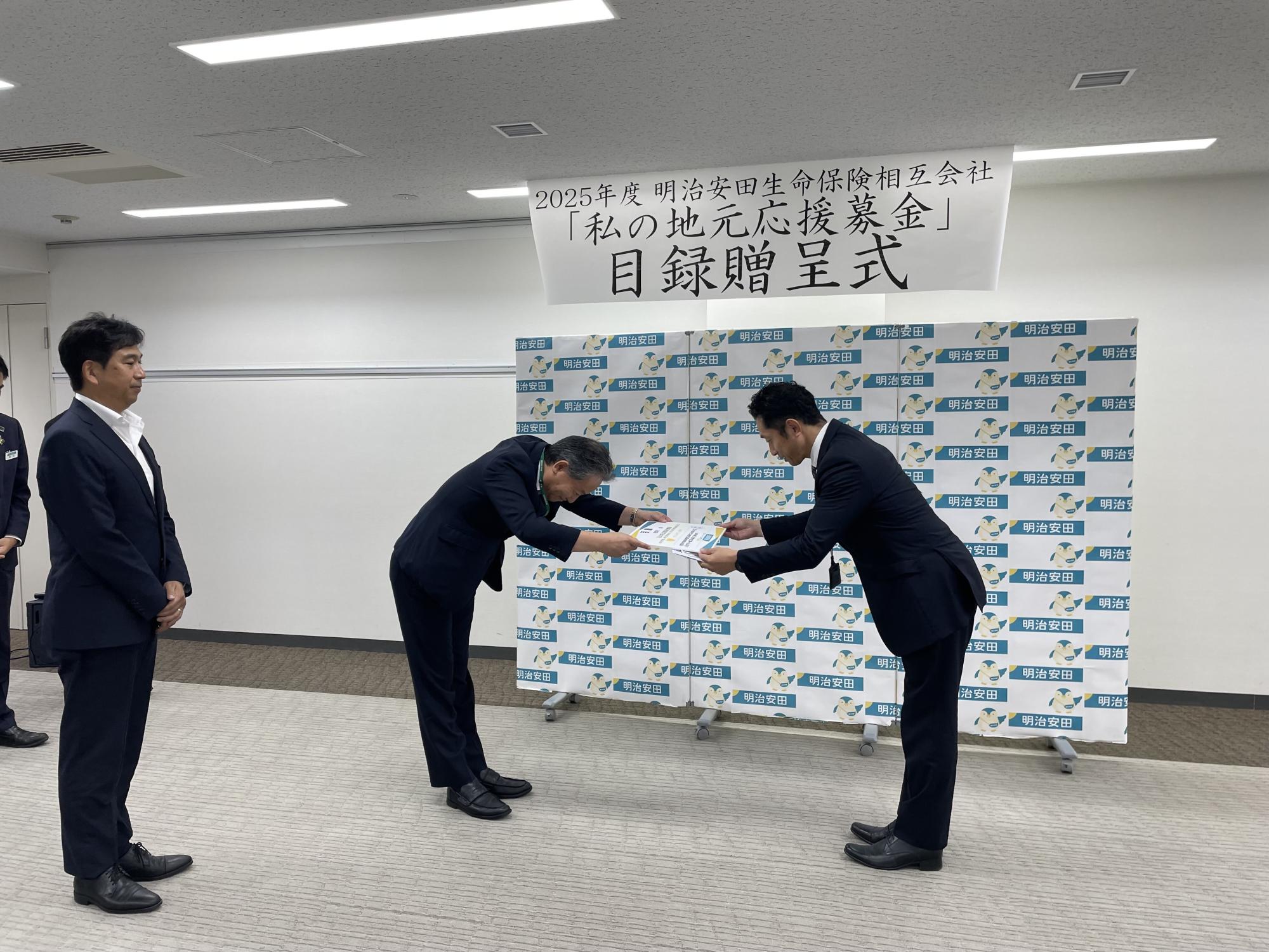 室内で「2025年度 明治安田生命保険相互会社 私の地元応援募金 目録贈呈式」と書かれた横断幕の前に、スーツ姿の二人が深くお辞儀をしながら書類を交換しており、背景には企業ロゴが並ぶパネルが設置された贈呈式の様子を示す写真
