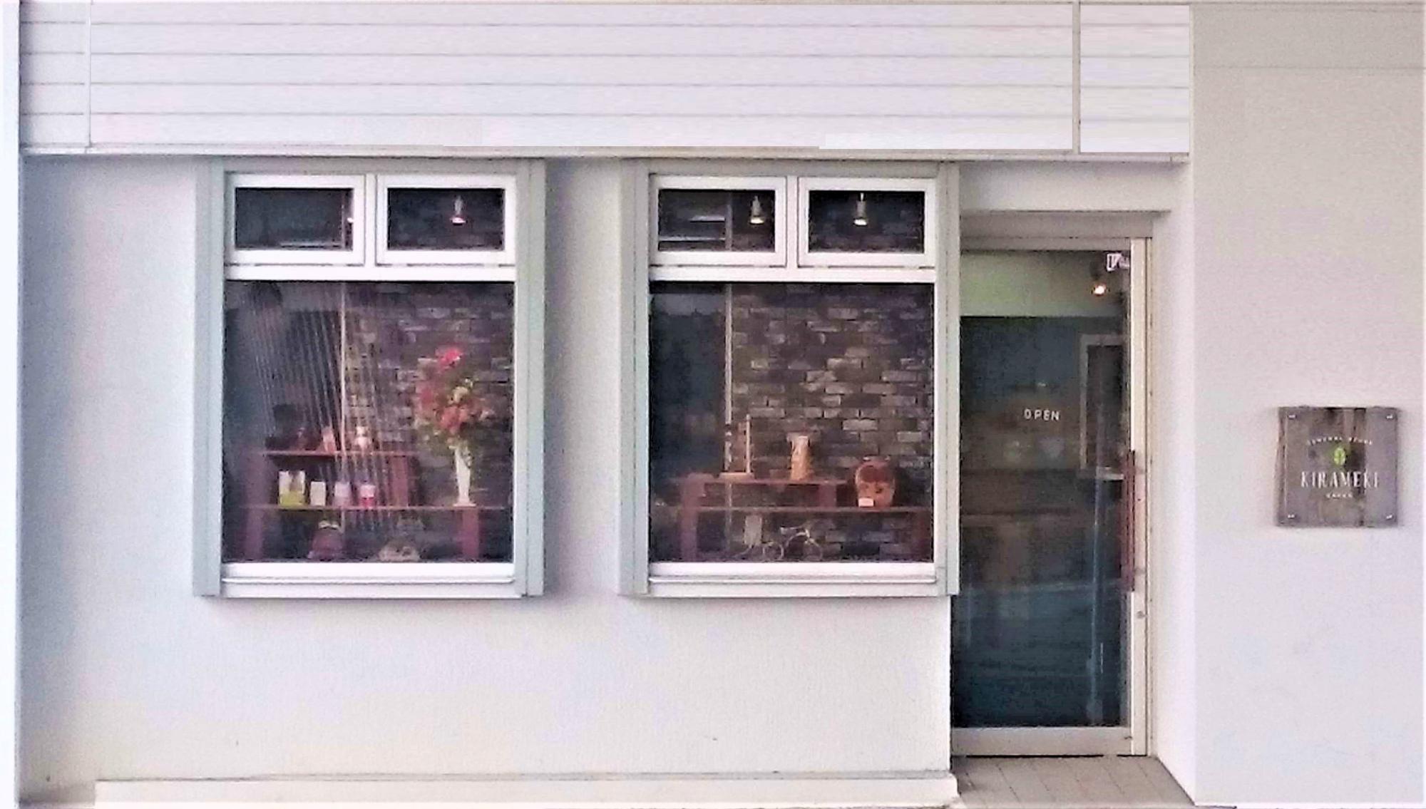 白い外壁に二つの大きな窓とガラス扉が並び、窓越しに店内の商品棚や装飾された花が見える「チャレンジドショップ きらめき」の外観写真