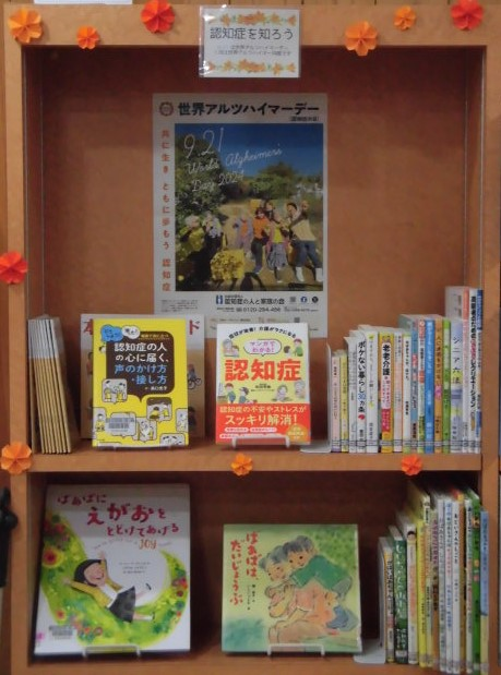 木製の本棚の枠にオレンジ色の花の飾りが付けられ、内部には「世界アルツハイマーデー」のポスターと共に、認知症への理解を深めるための書籍や絵本などが上下二段にわたって展示されている写真